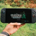 A hand holding a handheld gaming console displaying the Pokémon Legends game screen outdoors.